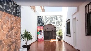 Un pasillo estrecho con una pared de piedra a la izquierda y una pared blanca a la derecha. Hay una planta en maceta a la izquierda y otra a la derecha. Un cartel con texto e imágenes es visible en el centro.
