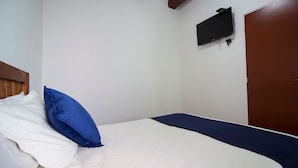 Una cama individual con ropa de cama blanca y azul, un cabecero de madera, un televisor de pantalla plana en la pared y una puerta marrón.