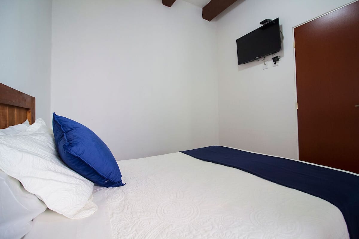 Una cama individual con ropa de cama blanca y azul, un cabecero de madera, un televisor de pantalla plana en la pared y una puerta marrón.