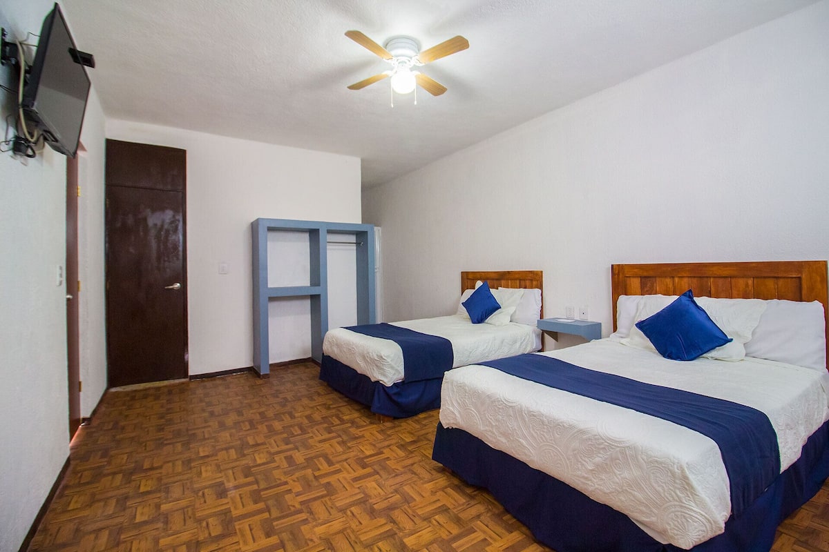 Habitación de hotel con dos camas, ventilador de techo, piso de madera y una mesita de noche.