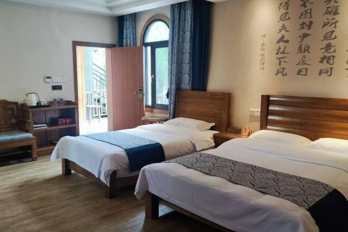 A hotel room with two beds, a wooden floor, a desk with a chair, and a window with curtains.