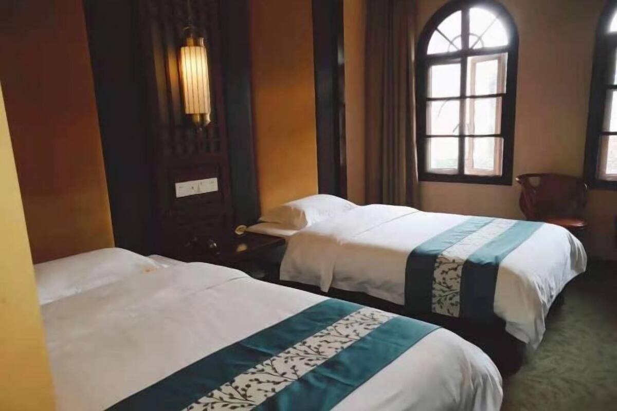 A hotel room with two beds, a nightstand, a lamp, and a window with curtains.