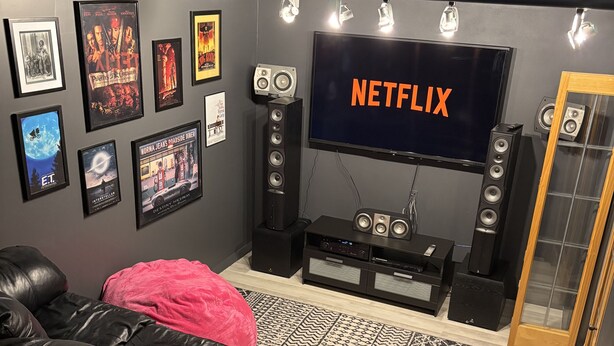 Un salon avec un canapé en cuir noir, un coussin rouge, un téléviseur à écran plat affichant le logo « NETFLIX », et un mur décoré d’affiches de films encadrées.