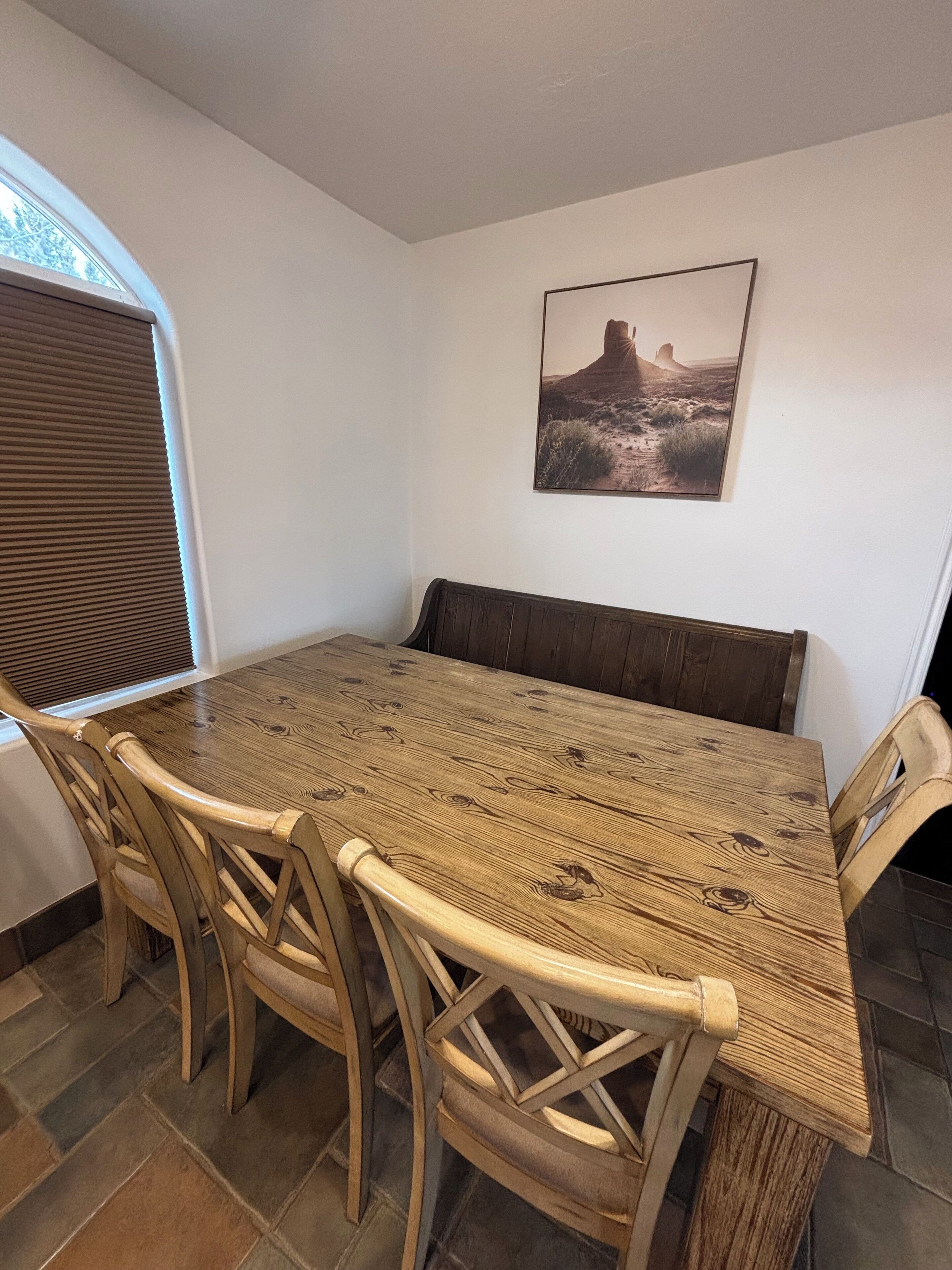 Un coin repas avec une table et des chaises en bois, un banc, une fenêtre avec des stores et un tableau accroché au mur.