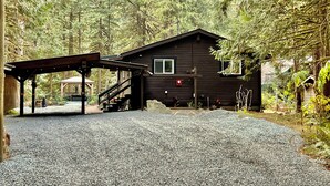 A rustic cabin with a gravel driveway, surrounded by dense forest. - Cozy Creekside Cottage (Hope)