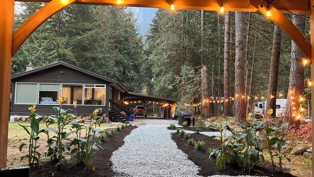 A cabin with a gravel pathway leading to a covered area with string lights. - Cozy Creekside Cottage (Hope)