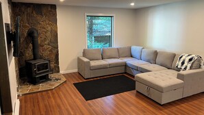 A modern living room with a stone wall, a sectional sofa, a black rug, and a wood-burning stove. - Cozy Creekside Cottage (Hope)