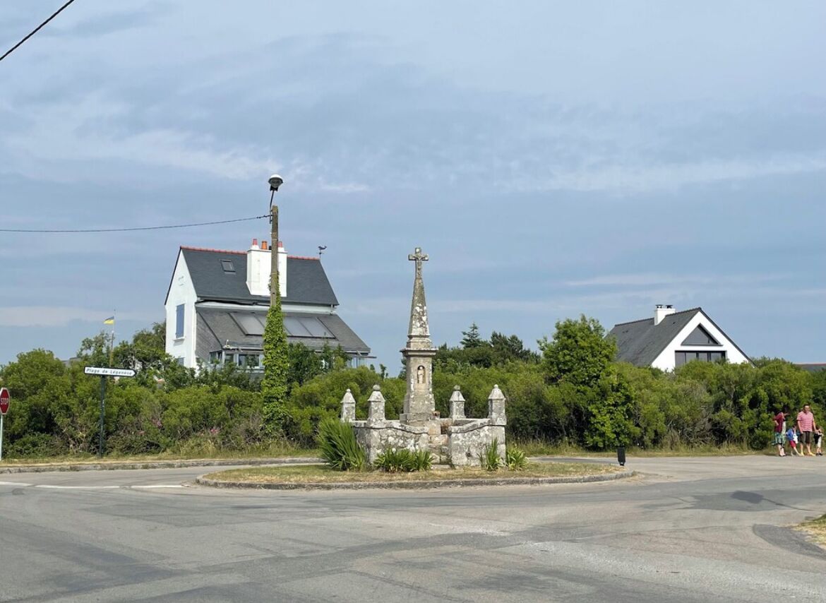 A roundabout with a stone monument in the center, surrounded by greenery and two houses with sloped roofs.