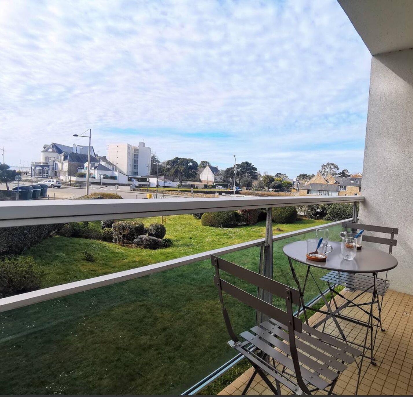 A balcony with a table and chairs, overlooking a grassy area and buildings in the distance.