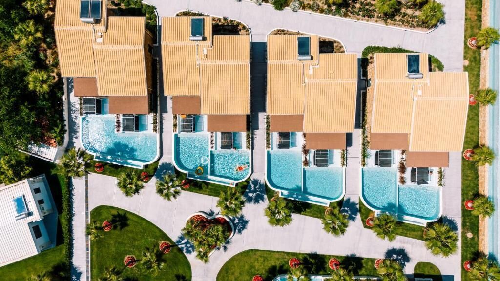 Una hilera de edificios modernos de dos pisos con piscinas en la azotea, rodeados de céspedes cuidados y palmeras.