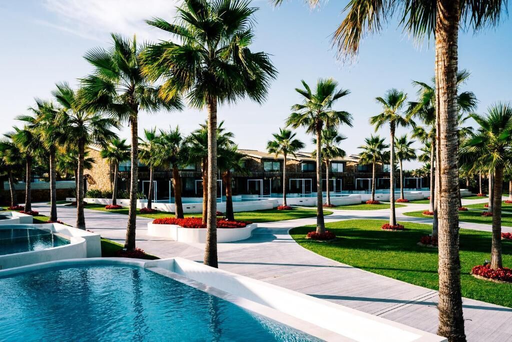 Un complex turístic amb piscina, palmeres i un edifici amb grans finestres.