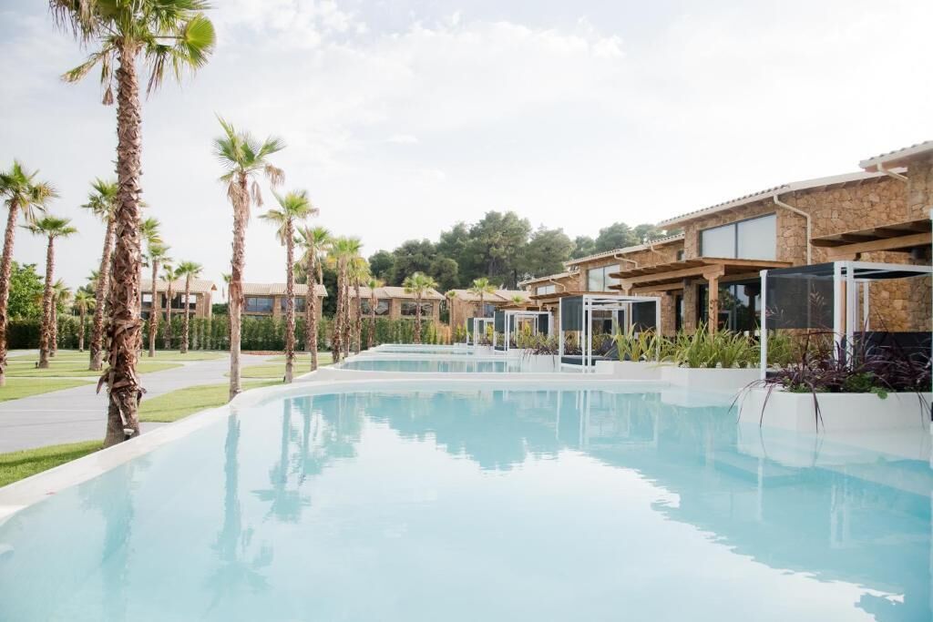 A swimming pool with a clear blue surface, surrounded by palm trees and modern buildings with stone facades.
