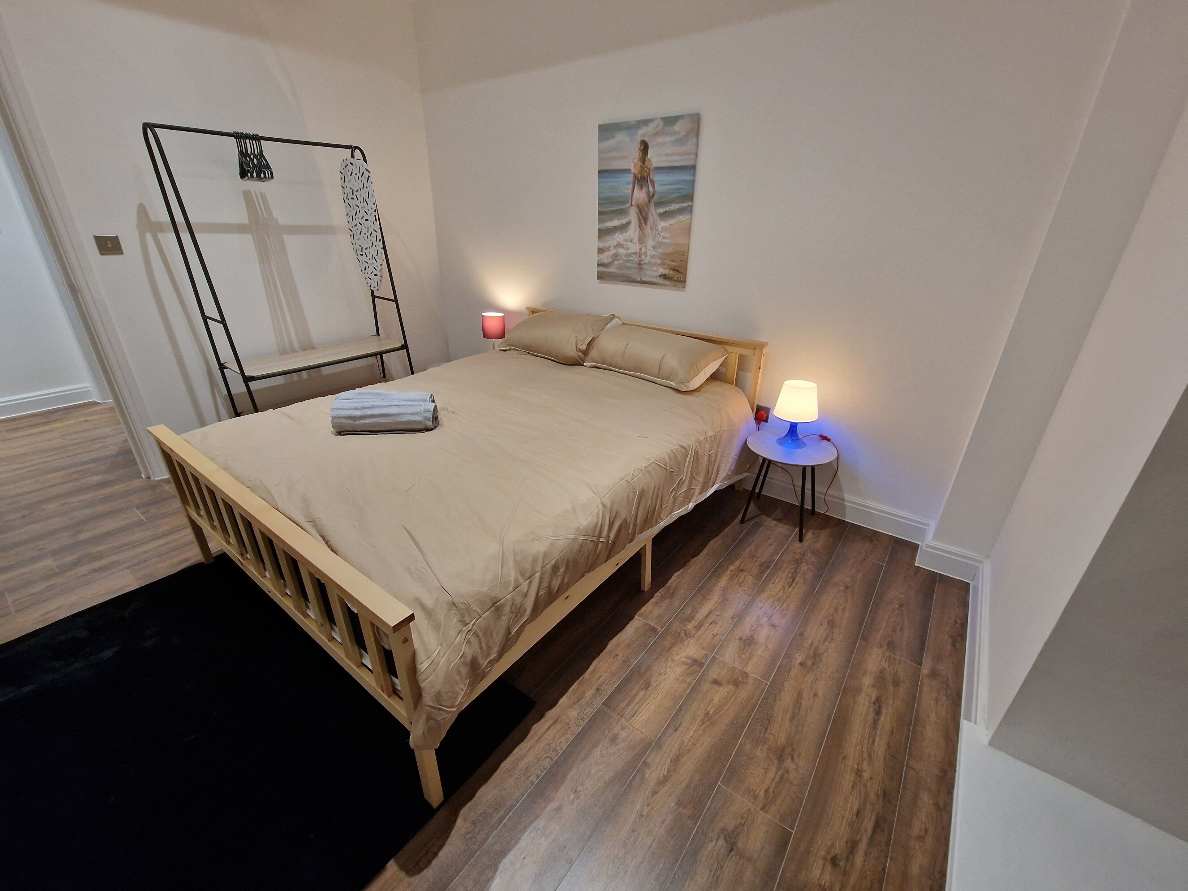 Een slaapkamer met een houten bed, een nachtkastje met een lamp, een wandplank en een schilderij van een strandgezicht.