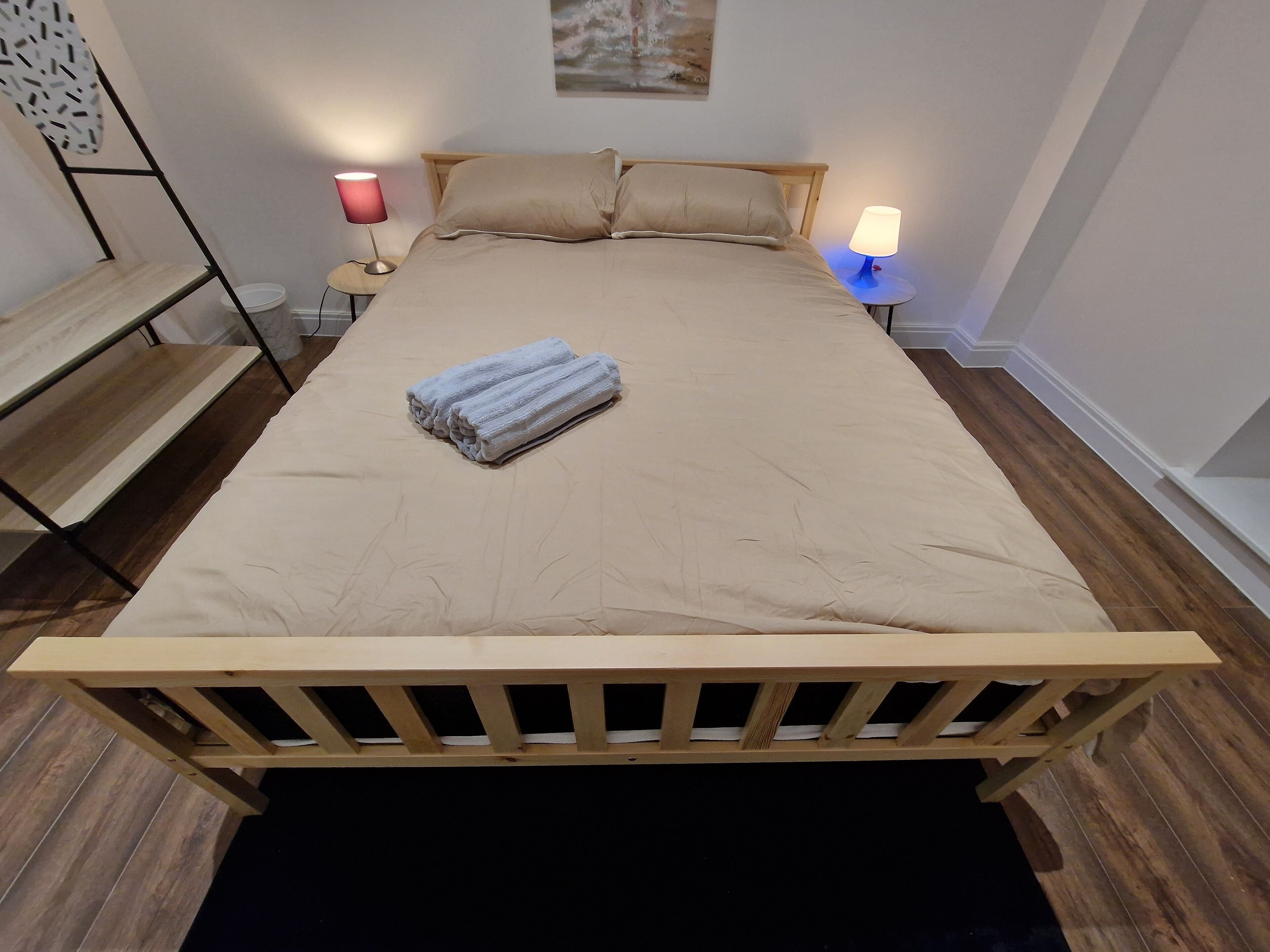 A neatly made bed with a beige bedspread and two pillows, a folded towel on the bed, and two bedside lamps on either side.