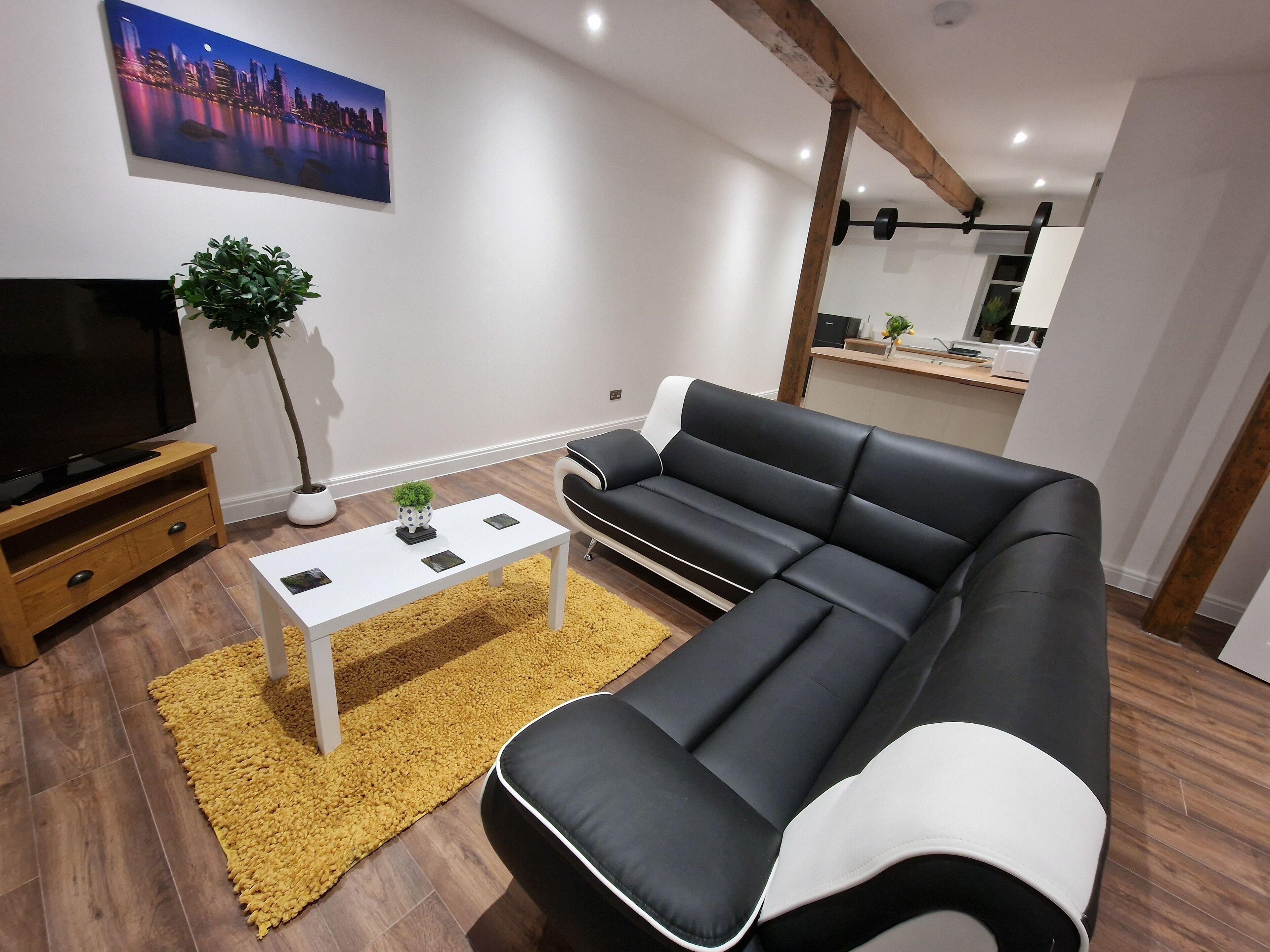 Una sala de estar moderna con un sofá seccional negro, una mesa de centro blanca, un televisor sobre un soporte de madera, una planta en maceta y una pintura con vista a un paisaje urbano.