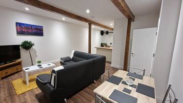 Een moderne woonkamer met een zwarte bank, een houten eettafel met zwarte placemats, een televisie op een houten dressoir, en een kleine plant.