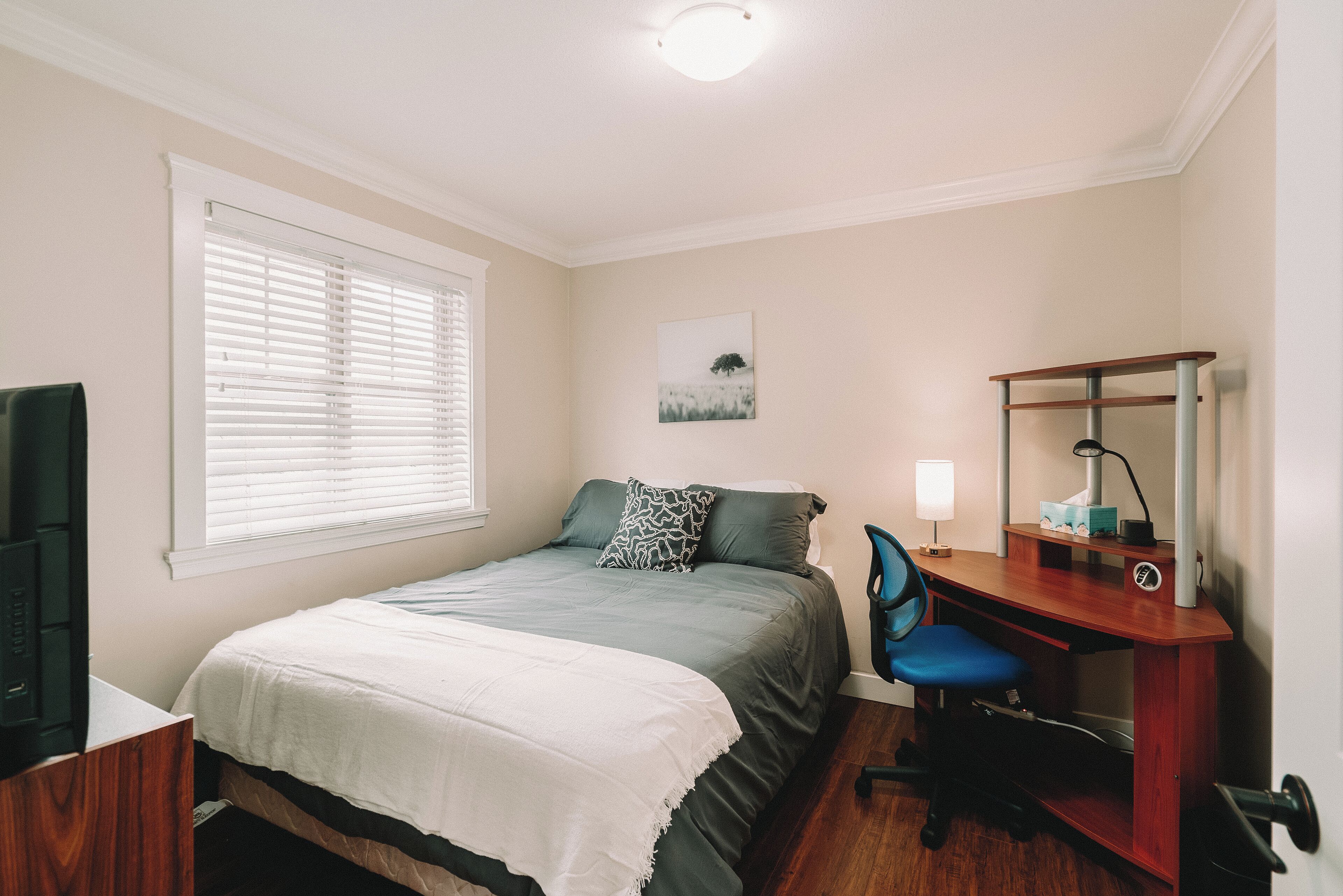 Une chambre à coucher comprenant un lit, un bureau, une chaise, un téléviseur et une fenêtre avec des stores.
