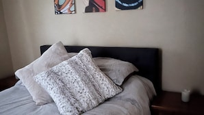 Uma cama com colcha cinza e duas almofadas, uma mesa de cabeceira com um vaso branco, e três quadros abstratos na parede.