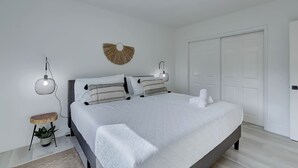 A modern bedroom with a large bed, a small side table with a plant, a wall-mounted light fixture, and a door.
