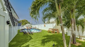 A backyard with a swimming pool, palm trees, and a white fence.