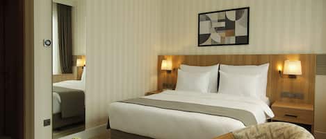 A hotel room with a large bed, bedside lamps, a wooden headboard, a small table with a cup and saucer, and a chair.