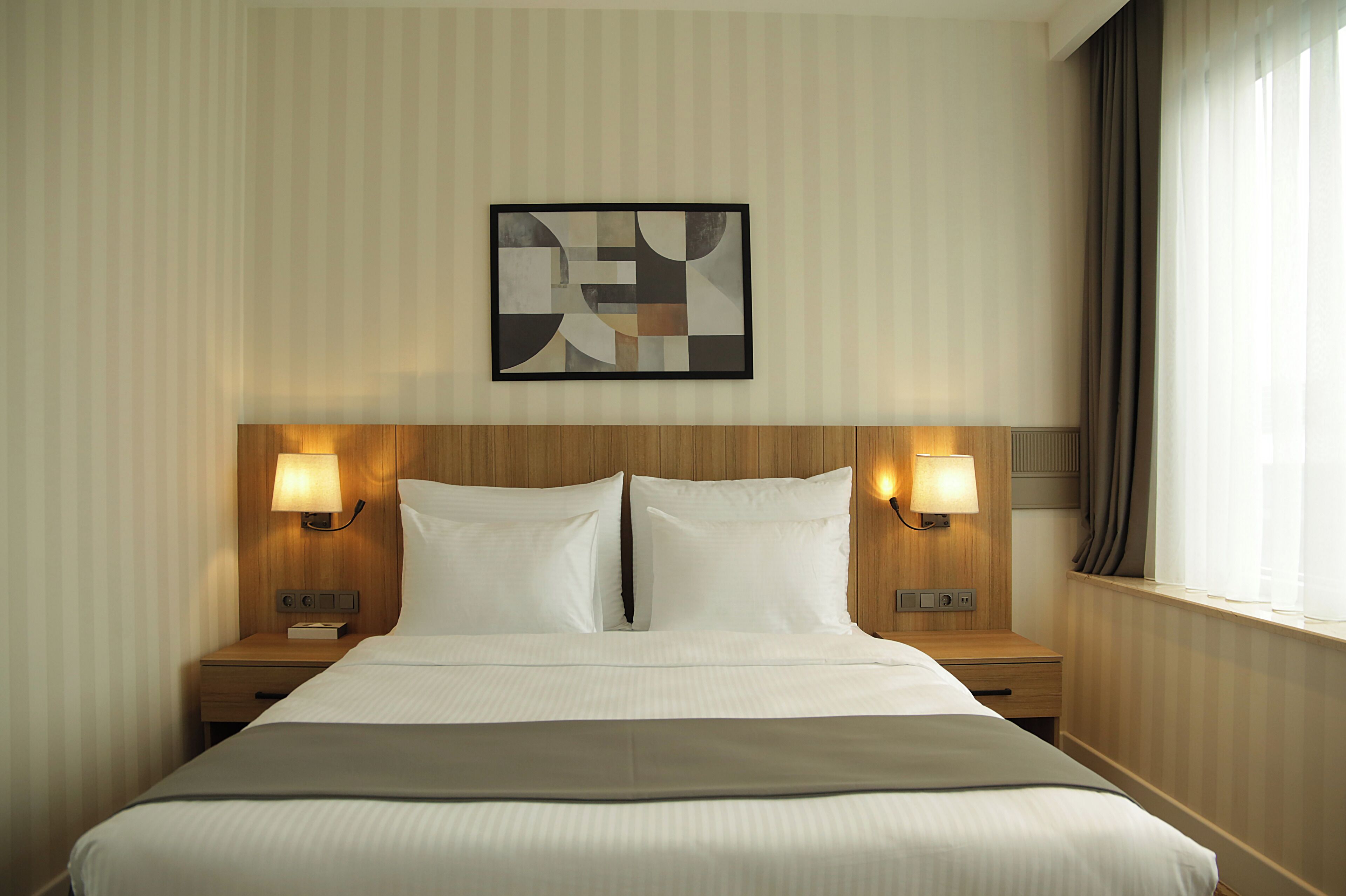 Ein Hotelzimmer mit einem großen Bett, zwei Nachttischen mit Lampen, einem gerahmten abstrakten Kunstwerk an der Wand und einem Fenster mit Vorhängen.