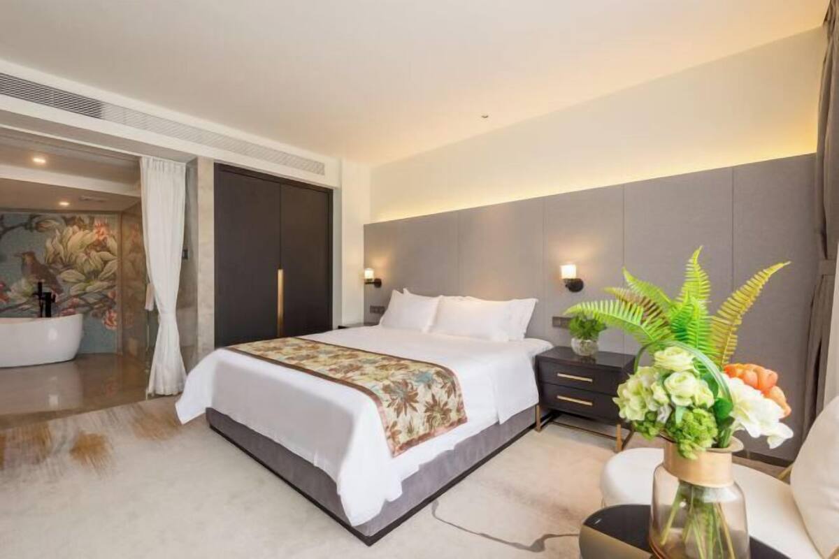 Une chambre d’hôtel moderne avec un grand lit, une table de chevet, un vase de fleurs et une salle de bain équipée d’une baignoire et d’une douche.