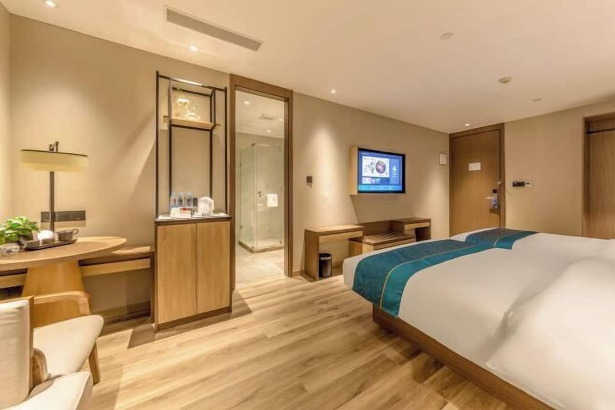 A modern hotel room with a bed, a desk, a chair, a TV, and a bathroom with a glass door.