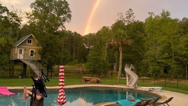 Een zwembad met een glijbaan, een klein huis op de achtergrond en een regenboog aan de hemel.