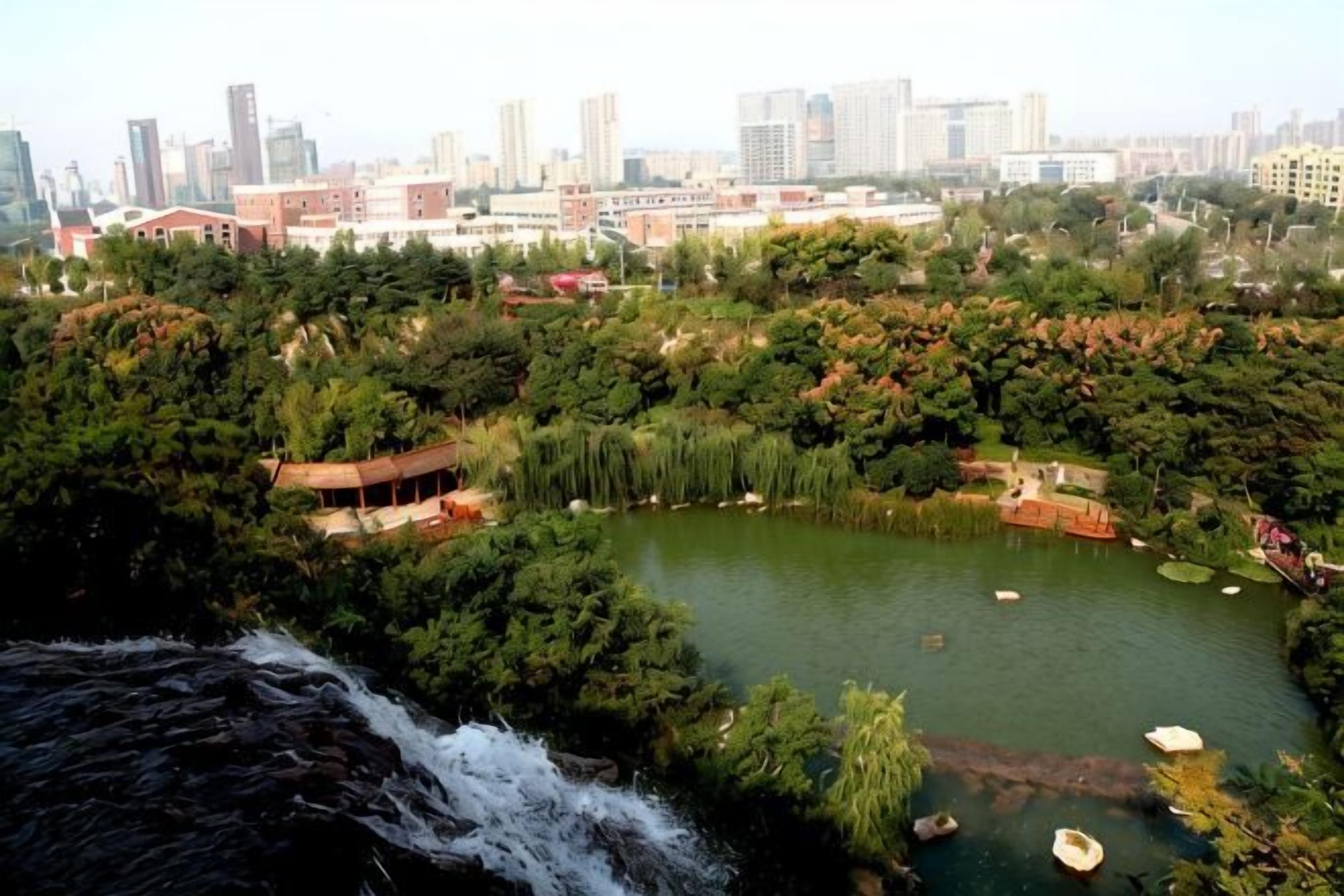 Uma cascata deságua em um lago rodeado por vegetação densa e uma paisagem urbana ao fundo.