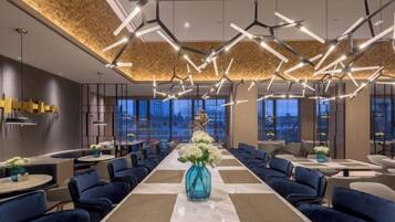 A modern hotel lobby with a long dining table, blue upholstered chairs, and contemporary lighting fixtures.