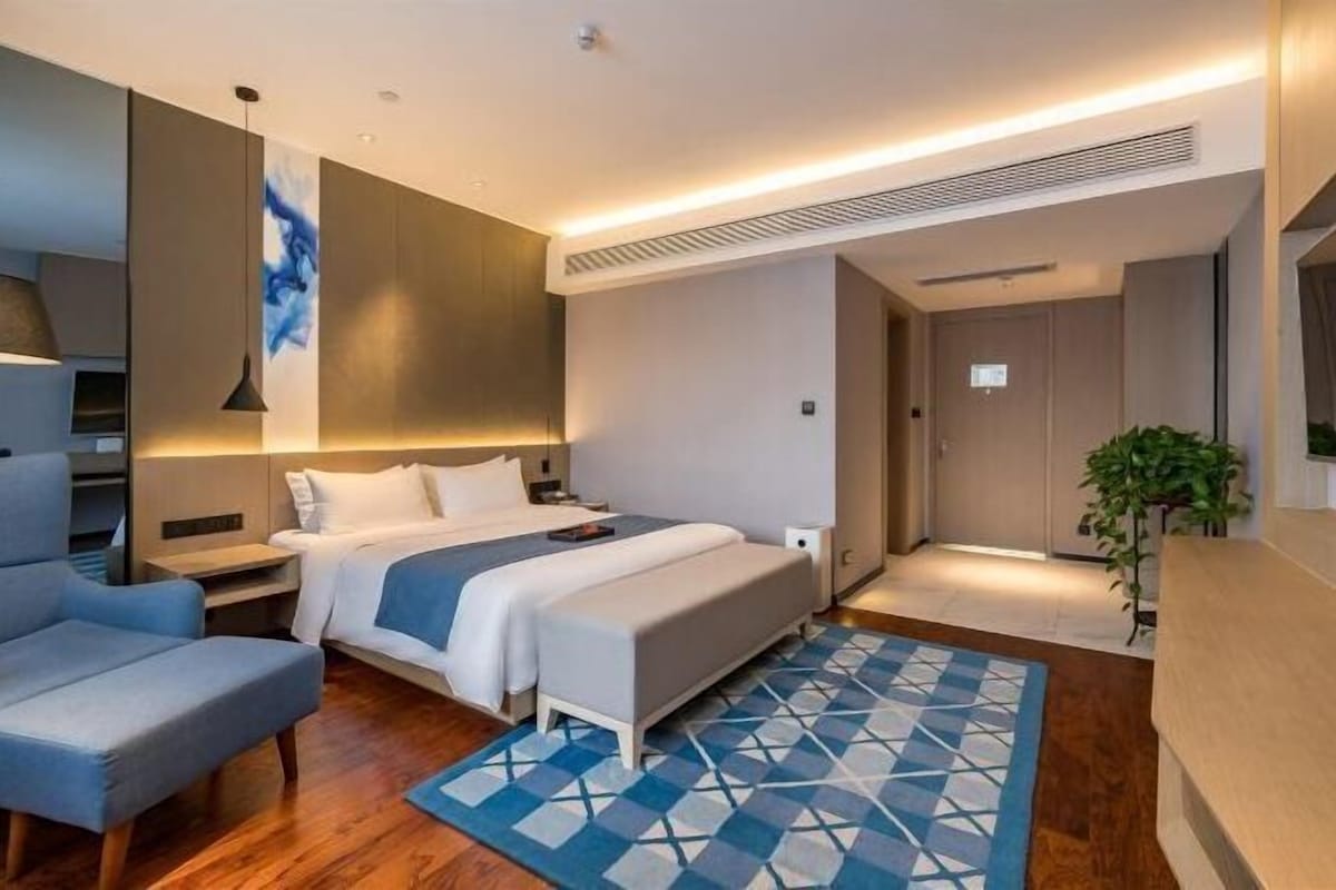 A modern hotel room with a bed, bedside tables, a chair, a plant, and a geometric-patterned rug.