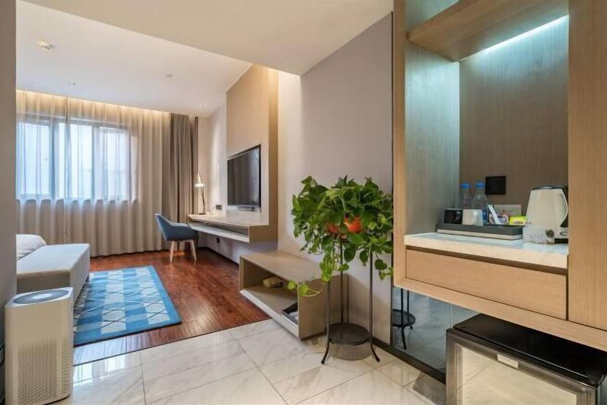 Une chambre d’hôtel moderne équipée d’un téléviseur à écran plat, d’un bureau, d’une chaise, d’une plante en pot, d’une machine à café et d’un petit coin cuisine.