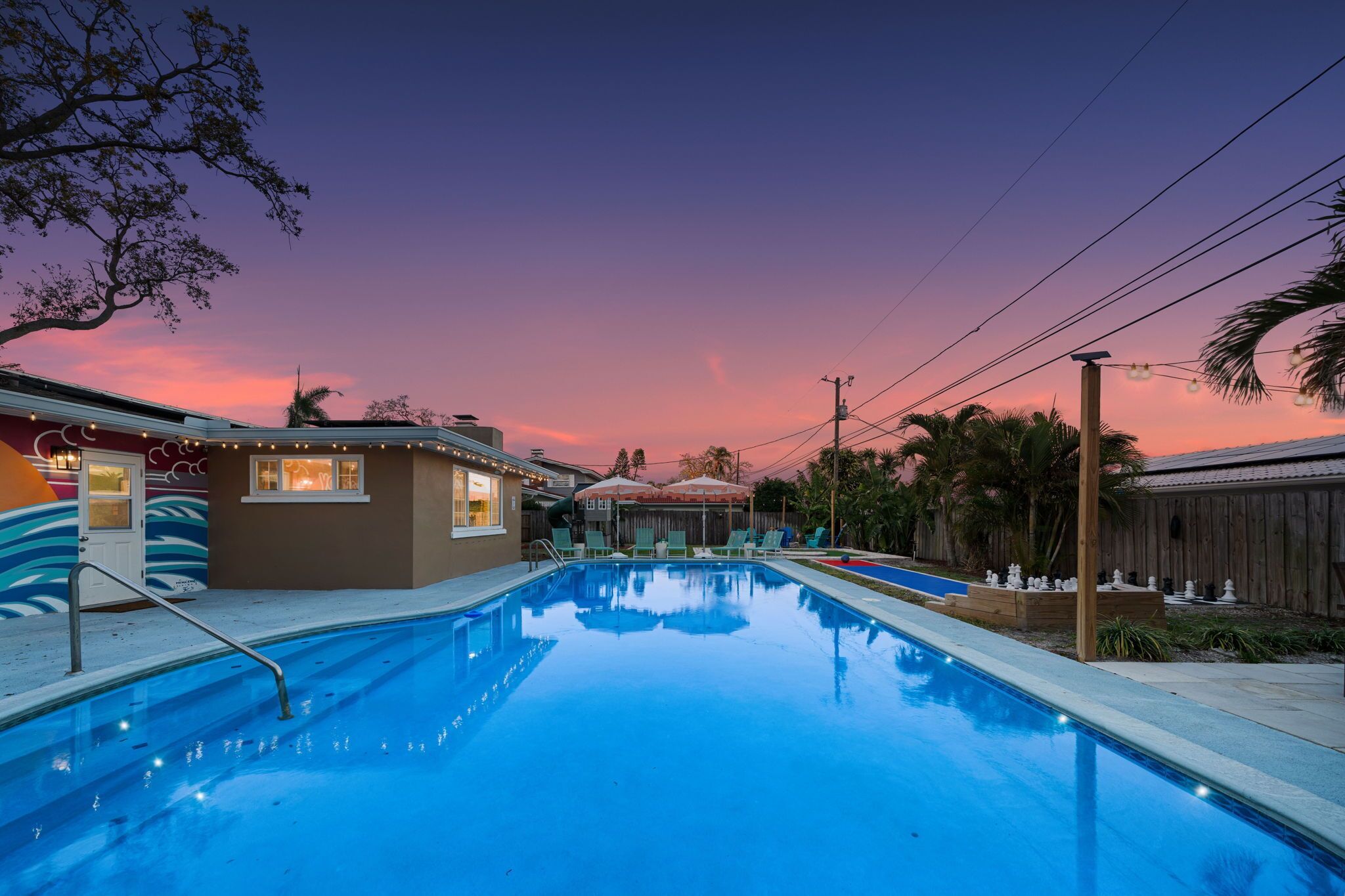 A swimming pool with lounge chairs and a wooden deck, surrounded by a wooden fence and palm trees.