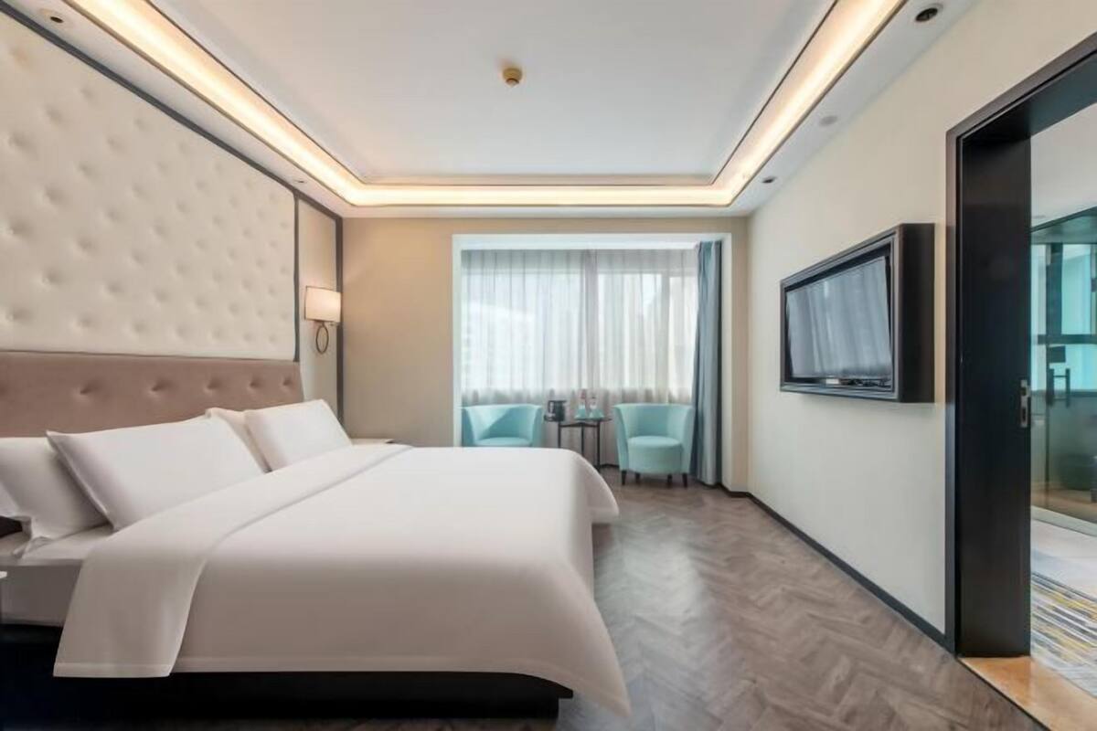 Habitación de hotel con una cama grande, un televisor, una zona de estar con dos sillones y un ventanal de vidrio que da a un balcón.