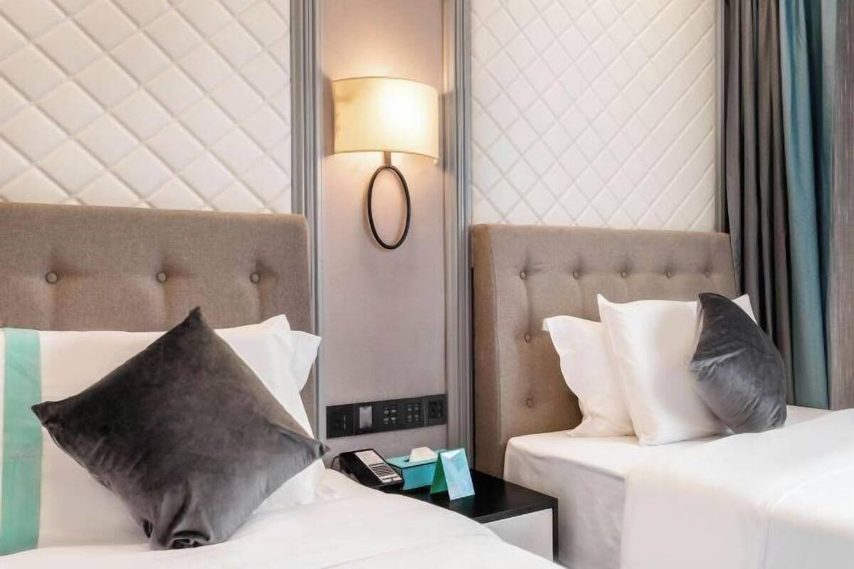 Habitación de hotel con dos camas, una lámpara de pared, una mesita de noche con teléfono y pañuelos, y un cabecero acolchado.