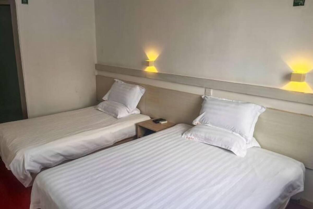 Habitación de hotel con dos camas individuales, cada una con ropa de cama y almohadas blancas, una mesita de noche con lámpara, y luminarias fijadas a la pared.