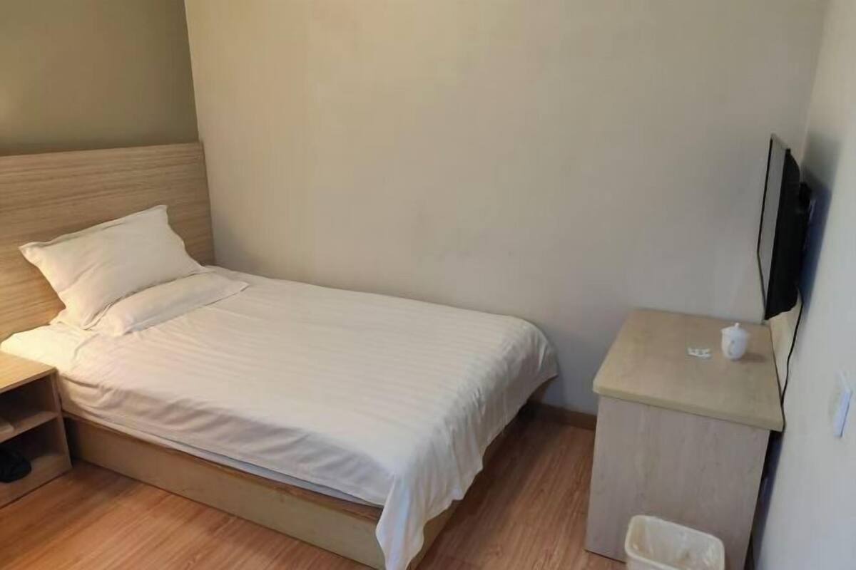 Una cama individual con cabecera de madera, una mesita de noche, un cubo de basura pequeño y un televisor montado en la pared.
