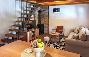 A modern living room with a wooden staircase, a black wood-burning stove, a sofa, a dining table with fruit, and a cowhide rug.