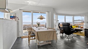 A spacious dining area with a view of a lake, a modern kitchen with large windows, and a cozy seating area with a yellow cushion. - 3 bedroom gorgeous home in Hejls (Hejls)