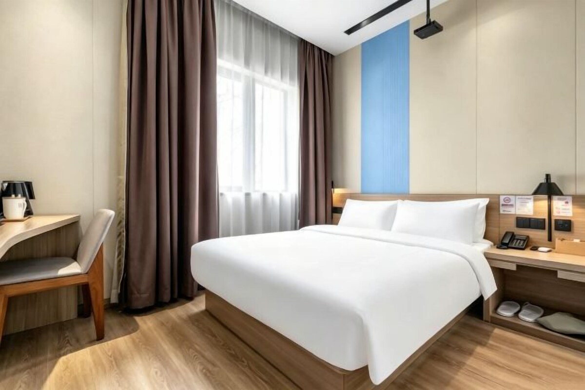 A hotel room with a bed, a desk, a chair, a window with curtains, and a nightstand with a lamp.