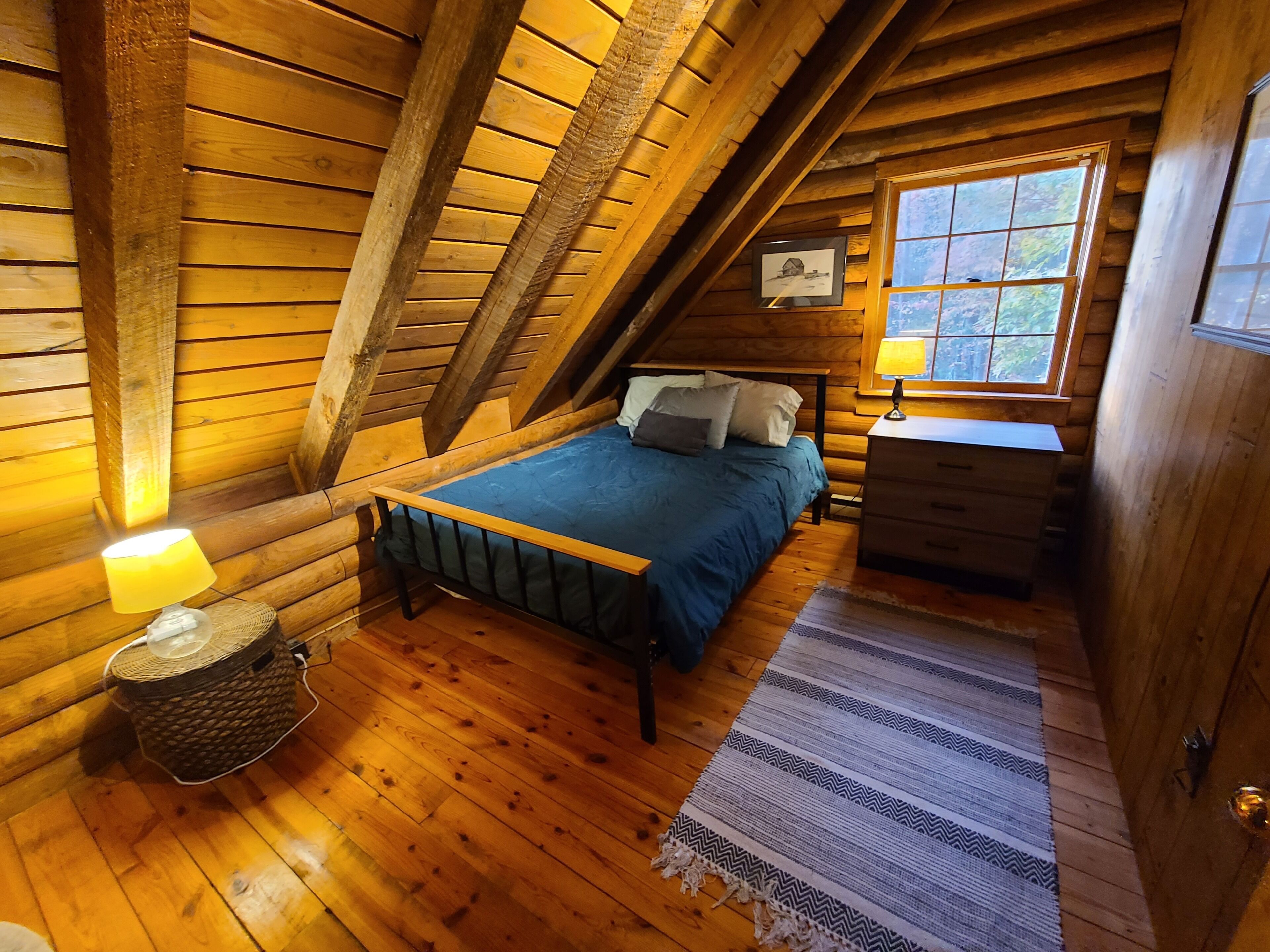 Une chambre confortable dans un chalet en bois, avec un plafond en bois, un lit avec une literie bleue, une table de chevet avec une lampe et une fenêtre donnant sur la forêt.