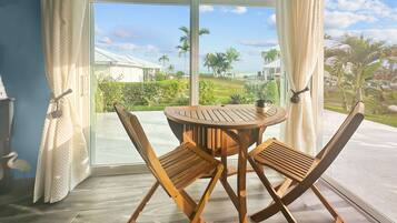 A room with a round wooden table and two wooden chairs, a large sliding glass door leading to a balcony with a view of a beach and palm trees.