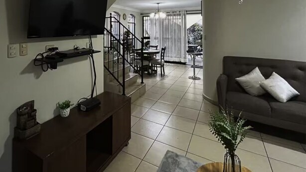 A living room with a flat-screen TV, a sofa, a coffee table, and a staircase leading to a dining area. - Residencia Cálida y acogedora (Santa Tecla)