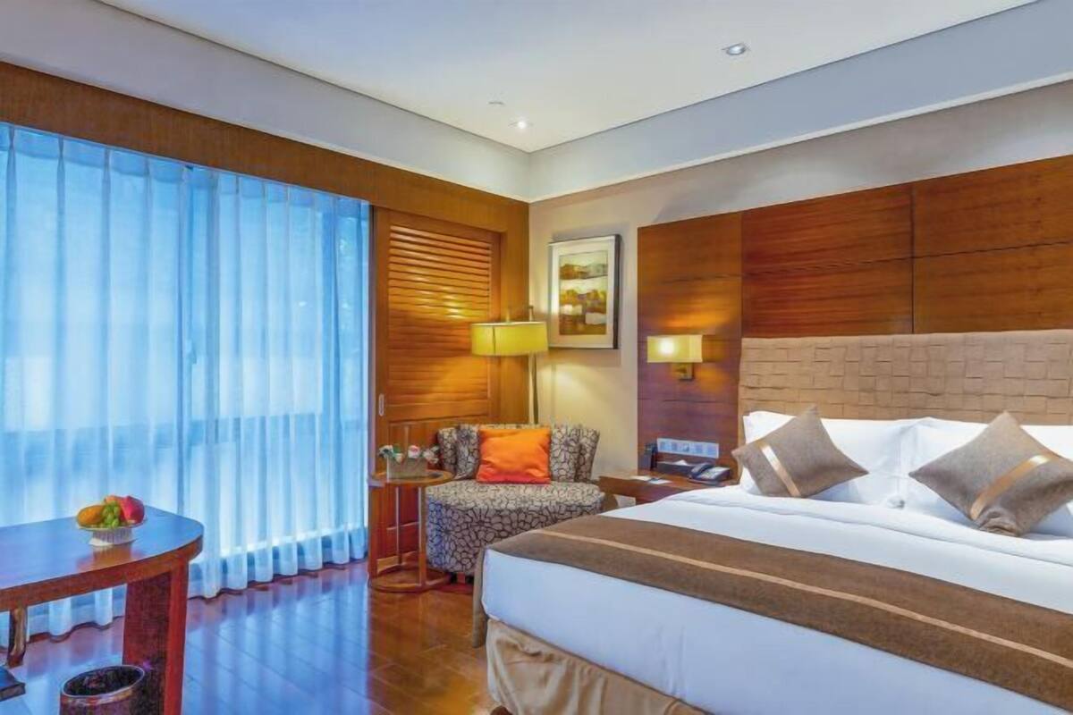 A hotel room with a bed, a small table with fruit, a chair, a lamp, and a painting on the wall.