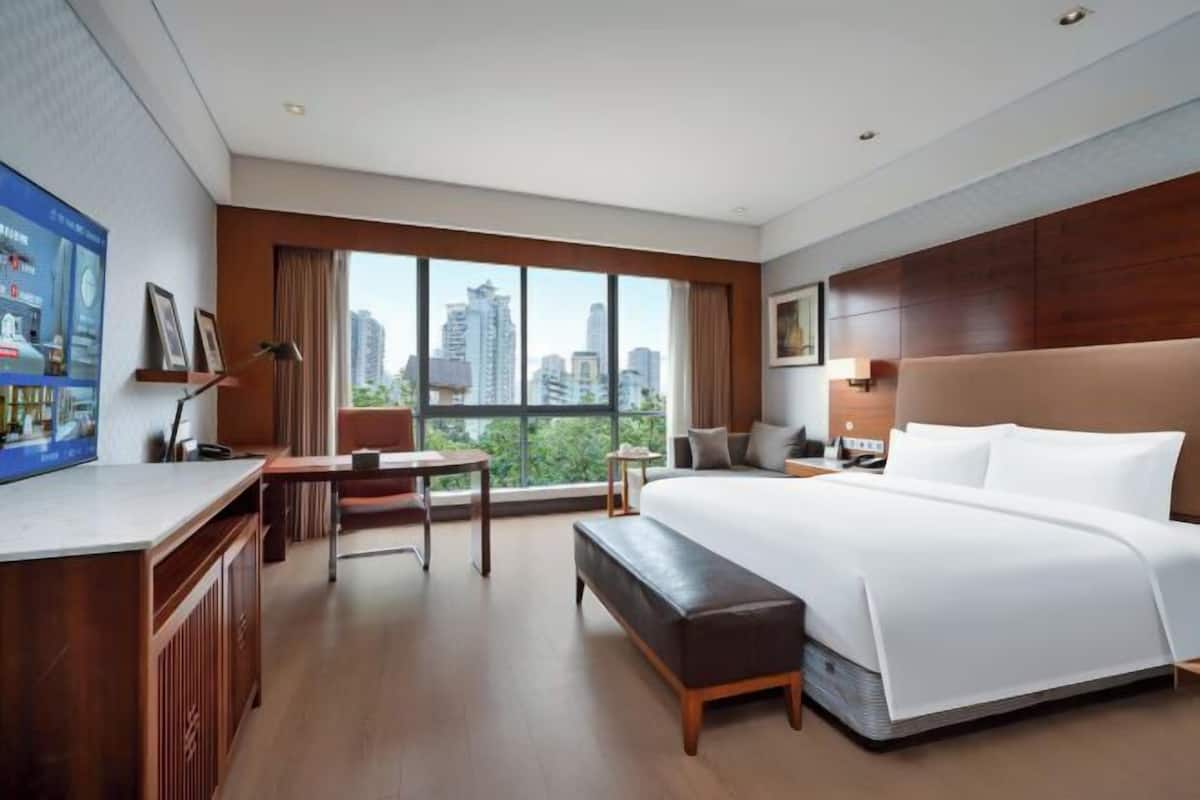 A modern hotel room with a large bed, a desk, a bench, and a television on the left wall.