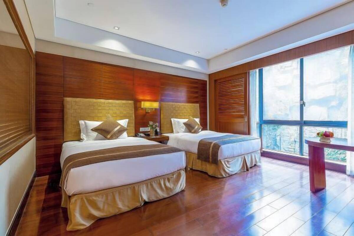 A hotel room with two beds, wooden paneling, a large window, and a small table.
