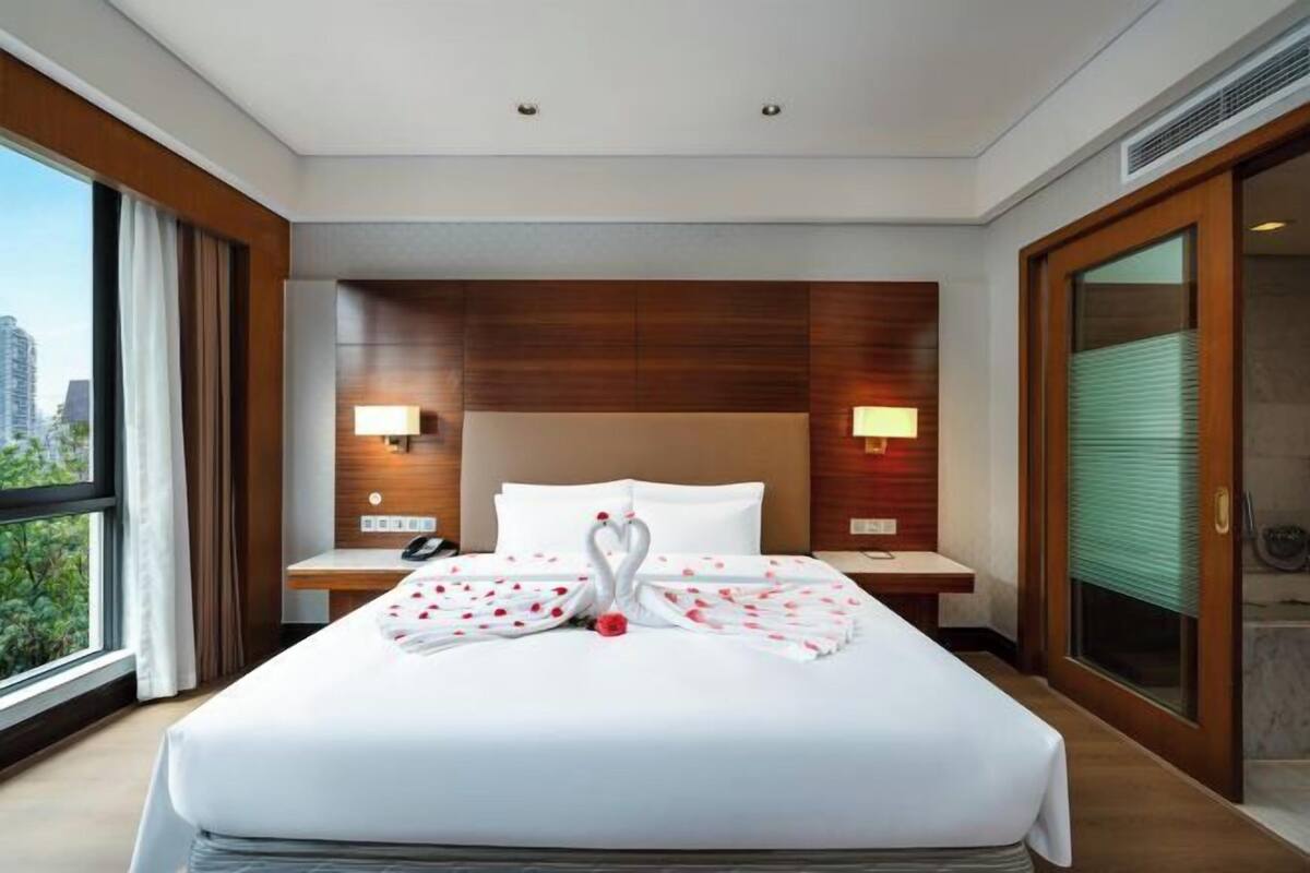 A hotel room with a large bed, two bedside lamps, a wooden headboard, and a view of the city.