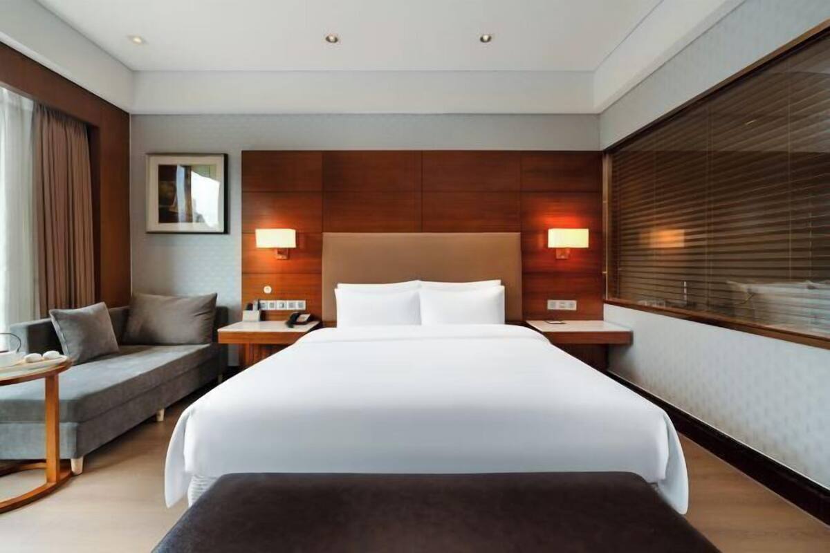A hotel room with a large bed, a sofa, a desk with a lamp, and a window with blinds.