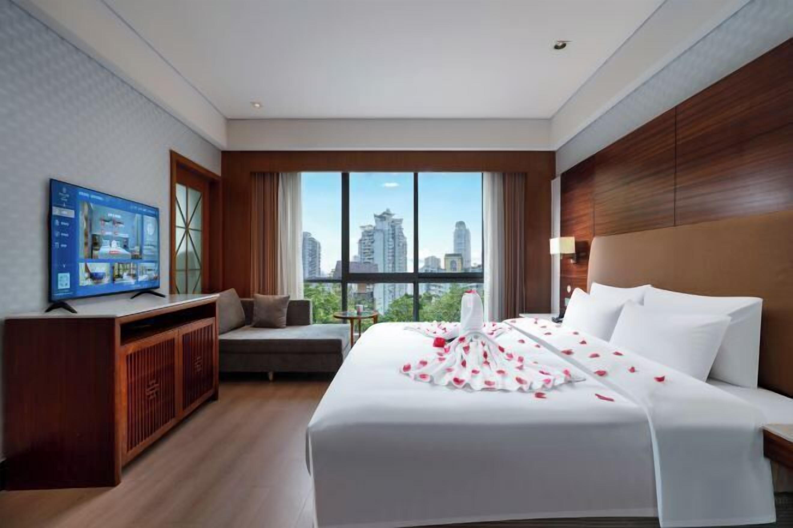 Quarto de hotel com uma cama grande, uma televisão sobre um armário de madeira, um sofá e vista para a cidade através de grandes janelas.